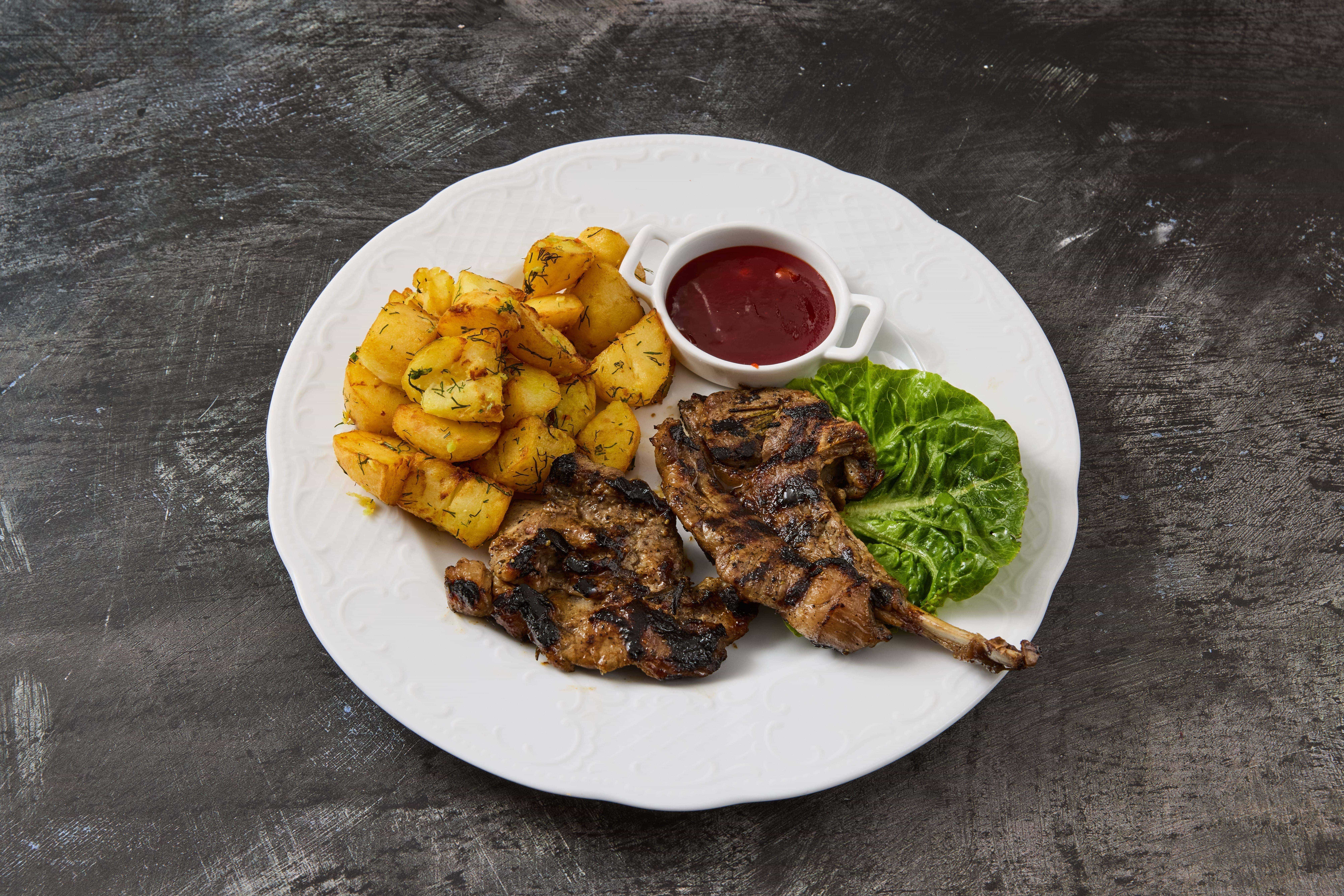 Gegrillte Lammkoteletts mit Ofenkartoffeln, frischem Salatblatt und hausgemachter Soße auf einem weißen Teller – Balkan Küche Spezialität