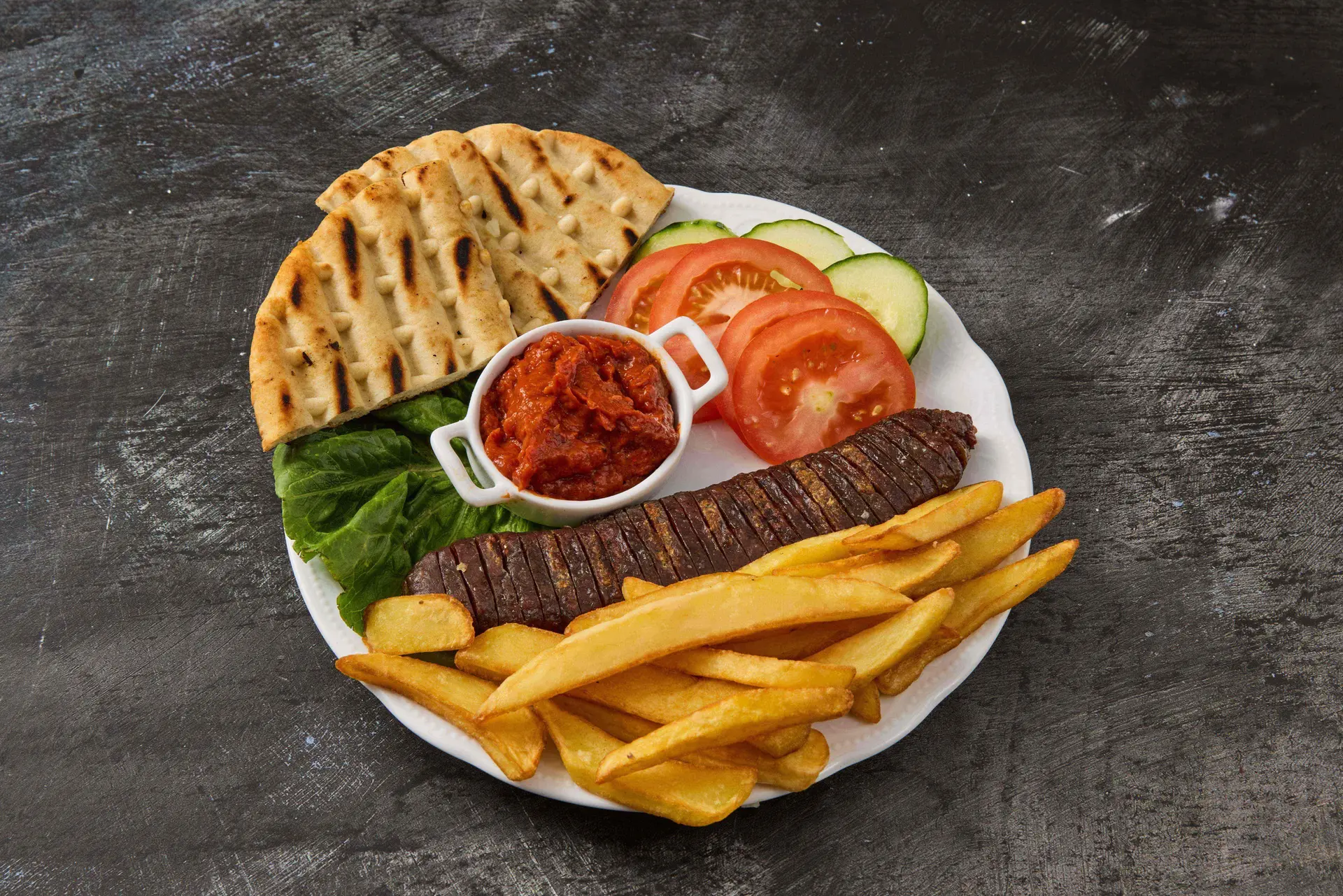 Teller mit gegrillter Rindswurst (Sucuk), Pommes frites, Ajvar, Fladenbrot, Tomaten- und Gurkenscheiben – traditionelles Balkan-Gericht